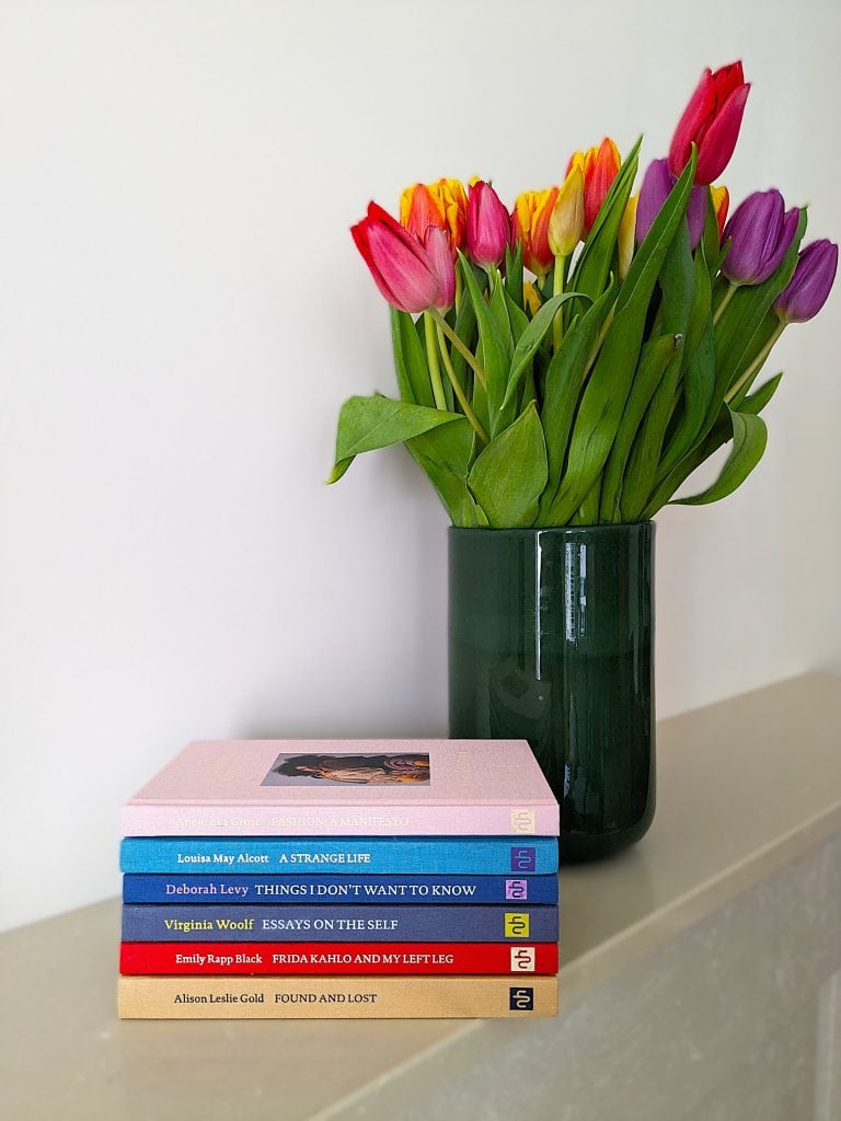 A stack of NHE books by women authors on a cream-coloured stone mantlepiece. A green vase full of colourful tulips stands next to the stack.